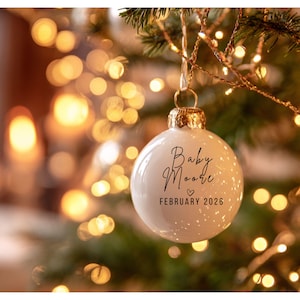 May include: White Christmas ornament with gold cap, hanging from a tree branch. The ornament has the text "Baby Moore" and "February 2026" in black script. The background is blurred with Christmas lights.