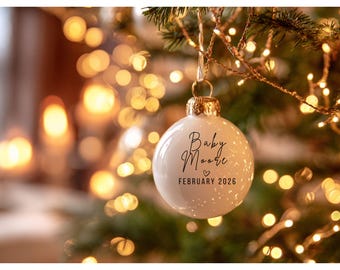 May include: White Christmas ornament with gold cap, hanging from a tree branch. The ornament has the text "Baby Moore" and "February 2026" in black script. The background is blurred with Christmas lights.