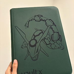 May include: A dark green Vault X trading card binder featuring a black graphic of a Rayquaza. The binder has a textured surface and the Vault X logo is embossed at the bottom. The binder is held by a hand.
