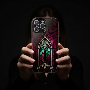 May include: A smartphone case featuring a stained-glass window design. The case has a dark red and magenta background with gold-colored trim. The stained-glass window design includes green and magenta glass pieces. The phone case has three camera lenses.