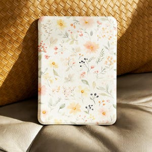 Puede incluir: Una funda rectangular con estampado floral. El diseño presenta flores de acuarela en amarillo, melocotón y rosa, con hojas verdes y bayas negras sobre un fondo crema. La funda está sobre un fondo neutro.