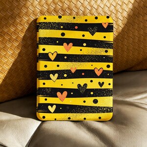 Pode incluir: Uma capa para tablet com um design de coração amarelo e preto. A capa apresenta listras pretas horizontais, listras amarelas e pontos pretos dispersos. Os corações são em tons de amarelo e laranja.