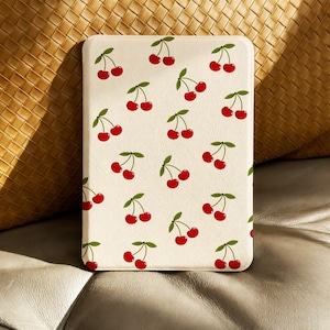 May include: A rectangular, cream-coloured case with a repeating pattern of red cherries and green leaves. The case is on a leather surface with a woven background.