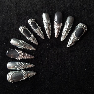Peut inclure: Un ensemble de dix faux ongles disposés en demi-cercle. Chaque ongle présente une forme pointue avec une finition argentée métallique, des motifs en relief complexes, et certains modèles incluent un panneau central noir mat avec des clous argentés.