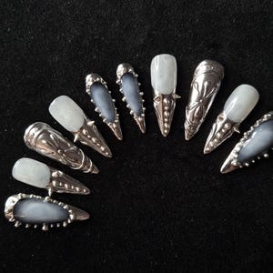 Op de afbeelding: Een set van tien press-on nagels in een puntige stiletto-vorm. De nagels hebben een mix van doorschijnende grijze en witte afwerkingen, geaccentueerd met ingewikkelde zilverkleurige metalen versieringen, waaronder studs, kralen en bloemmotieven.