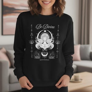 May include: Black sweatshirt with a white graphic design. The design features the text "Be Divine" above a woman's face, a crescent moon, crystals, and other celestial elements. The sweatshirt is a crew neck style.