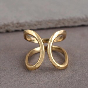 Puede incluir: Un anillo de tono dorado con un diseño abierto de estilo bypass, que presenta dos bucles redondeados y simétricos que se encuentran en el centro. El metal tiene un acabado liso y pulido, creando una forma fluida alrededor del dedo.