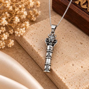 Puede incluir: Un collar con colgante de metal plateado que presenta un diseño cilíndrico con la cabeza detallada de una criatura con cuernos en la parte superior. El colgante cuelga de una cadena fina, mostrando intrincados patrones grabados y un acabado metálico pulido.