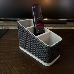 May include: A black and gray remote control holder with three compartments. The holder has a geometric pattern on the outside and a white trim. A black remote control is placed inside the holder.