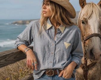 May include: A light blue button-down shirt with cream-colored lace accents on the collar and chest pocket, paired with a brown leather belt, blue jeans, and a straw cowboy hat, standing next to a horse with a brown leather bridle and reins.