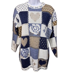 Könnte beinhalten: Ein Patchwork-Pullover in Blau-, Beige- und Weißtönen. Der Pullover zeigt verschiedene Muster, darunter Herzen, Rosen und geometrische Formen. Der langärmlige Pullover hat einen Rundhalsausschnitt und eine lockere Passform.