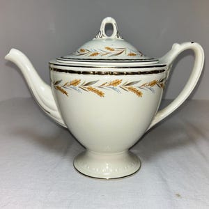 May include: Cream-colored ceramic teapot with a lid, featuring a wheat stalk design in gold, brown, and blue. The teapot has a curved spout, a handle, and a pedestal base. The lid has a loop handle.