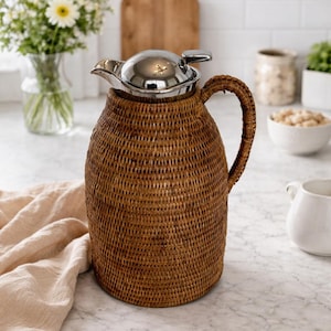 Puede incluir: Jarra de ratán tejido marrón con tapa y asa de acero inoxidable. La jarra tiene forma cilíndrica y está sobre una encimera de mármol. Probablemente se utiliza para servir bebidas. Altura aproximada 25 cm.
