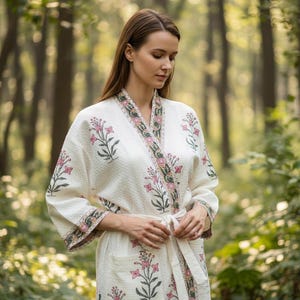 May include: White waffle-weave robe with a floral pattern in pink and green. The robe has a shawl collar, pockets, and a matching tie belt. The floral design is repeated on the cuffs and along the edges of the robe.