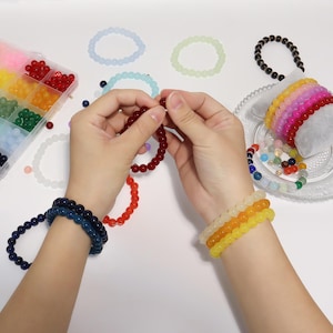 May include: A variety of colorful beaded bracelets in shades of red, blue, yellow, orange, and green. The image also shows a tray of beads in a divided container, and a person's hands assembling a bracelet.