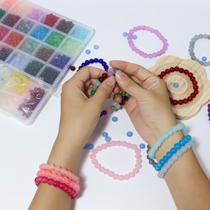 May include: A collection of colorful beaded bracelets and loose beads. A clear plastic organizer holds various shades of beads. Several bracelets are worn on wrists, including pink, blue, and gray. The image showcases the beads and bracelets.