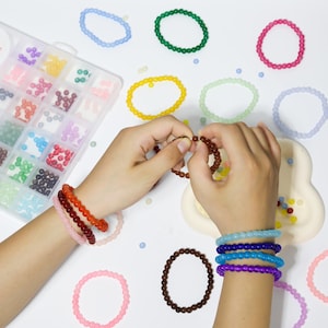 May include: A collection of colorful beaded bracelets in various shades of pink, orange, brown, blue, and purple. The image shows a person's hands assembling a bracelet, with a clear plastic organizer filled with beads in the background.
