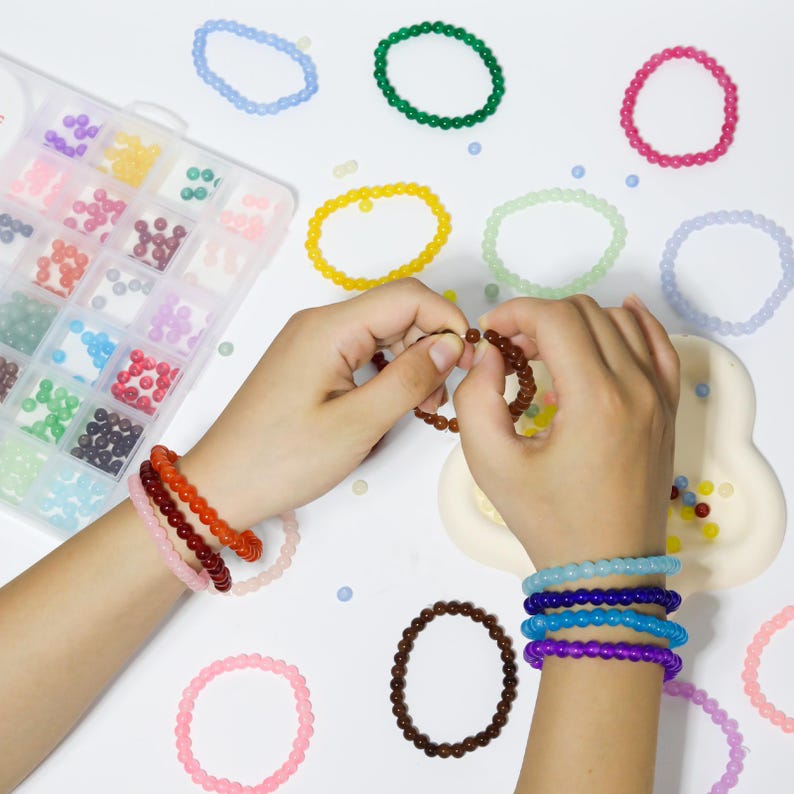 May include: A collection of colorful beaded bracelets in various shades of pink, orange, brown, blue, and purple. The image shows a person's hands assembling a bracelet, with a clear plastic organizer filled with beads in the background.