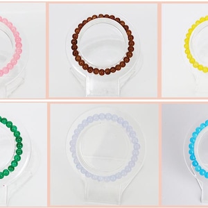 May include: Six beaded bracelets in various colors: pink, brown, yellow, green, lavender, and blue. Each bracelet is displayed on a clear, circular stand, showcasing the round beads. The bracelets are likely for wrist wear.