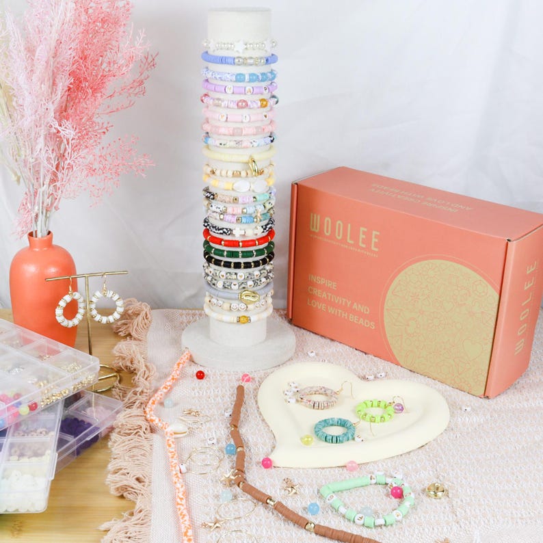 May include: A display of colorful bracelets on a white stand, with a coral-colored box that reads "WOOLEE" and "INSPIRE CREATIVITY AND LOVE WITH BEADS." A vase with pink flowers and a pair of earrings are also visible. A heart-shaped tray holds more bracelets.