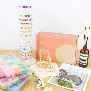 May include: A display of colorful beaded bracelets on a white stand, alongside a box labeled "WOOLEE" and containers of beads. A pearl necklace rests on a cloud-shaped dish. The scene is set on a light-colored wooden surface.