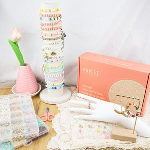 May include: A display of colorful beaded bracelets on a white stand, alongside a jewelry-making kit and various earrings. The bracelets feature a variety of colors and designs. A pink vase with a tulip and a pair of pink scissors are also visible.