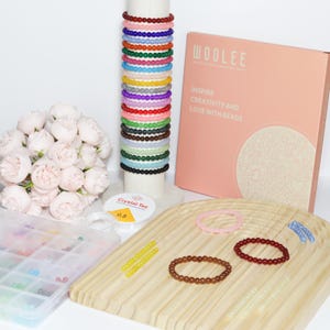 May include: A collection of colorful beaded bracelets in various colors, stacked on a white cylinder. A pink box with the text "WOOLEE" and "INSPIRE CREATIVITY AND LOVE WITH BEADS" is visible. Additional bracelets and beading supplies are also present.