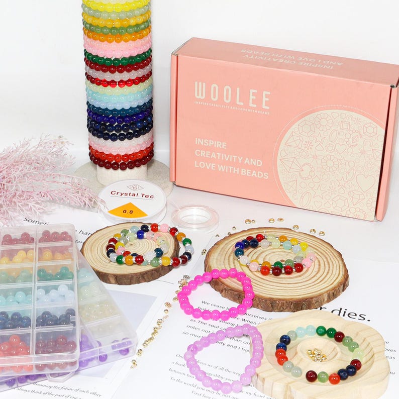 May include: A collection of colorful beaded bracelets and jewelry-making supplies. The image includes a pink box with the text "WOOLEE" and "INSPIRE CREATIVITY AND LOVE WITH BEADS". There are also spools of string and a container of beads.