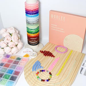 May include: A collection of colorful beads and bracelets, including a display of stacked bracelets in various colors. A pink box with the text "WOOLEE" and "INSPIRE CREATIVITY AND LOVE WITH BEADS" is also present, along with a container of beads and string.