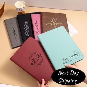 May include: A collection of personalized leather portfolios in various colors, including gray, black, pink, brown, and teal. Each portfolio features custom text, such as names and phrases, in elegant script. The portfolios are arranged on a light-colored surface.