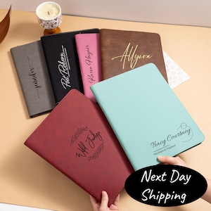 May include: A selection of personalised leather portfolios in shades of grey, black, pink, brown, burgundy, and teal. Each portfolio displays custom text, including names and phrases, in an elegant script. The portfolios are arranged on a light-coloured surface.