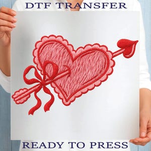 May include: A red heart design with a scalloped edge and an arrow piercing through it. A red bow is tied around the arrow. The words "DTF TRANSFER" and "READY TO PRESS" are printed on the image.