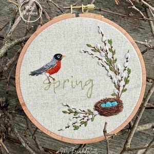 May include: Embroidered spring-themed hoop art featuring a robin, nest with blue eggs, and the word "spring". The design includes green leaves and white buds. The hoop is set against a background of twigs.
