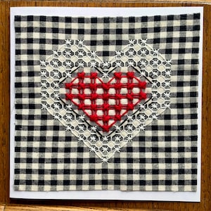 May include: A handmade greeting card featuring a red stitched heart on a white and black gingham background. The heart is surrounded by a white lace-like design. The card is square and set against a wooden surface.