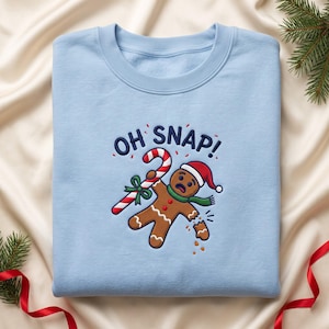 May include: A light blue crewneck sweatshirt featuring an embroidered gingerbread person wearing a red Santa hat and green scarf. The gingerbread person holds a red and white candy cane, with the text OH SNAP! embroidered above.
