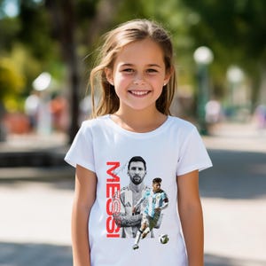 May include: White t-shirt featuring a graphic of a soccer player with the name "MESSI" in red vertical text. The design includes two images of the player in action. The shirt is worn by a young girl.