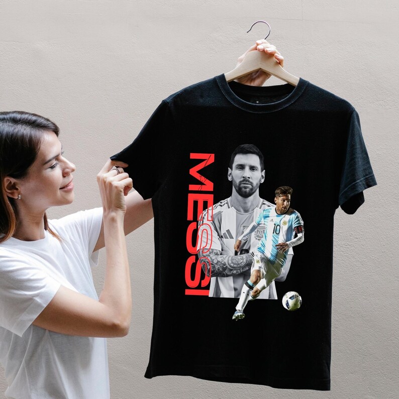 May include: Black t-shirt featuring a portrait of a soccer player with the name "MESSI" in red vertical text. The shirt displays two images of the player in action, one in a light blue and white uniform, and the other in a black and white portrait.