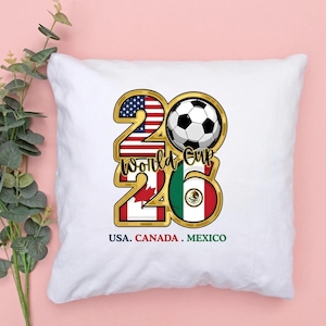 May include: White square pillow with a gold-outlined design featuring a soccer ball and the text "2026 World Cup." The design incorporates the flags of the USA, Canada, and Mexico. The text "USA. CANADA. MEXICO" is at the bottom.