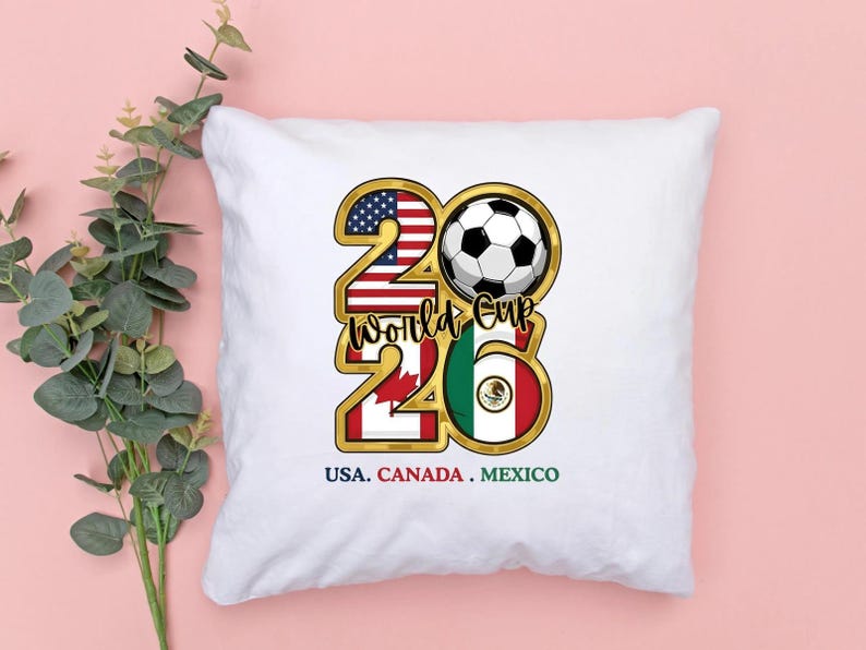 May include: White square pillow with a gold-outlined design featuring a soccer ball and the text "2026 World Cup." The design incorporates the flags of the USA, Canada, and Mexico. The text "USA. CANADA. MEXICO" is at the bottom.
