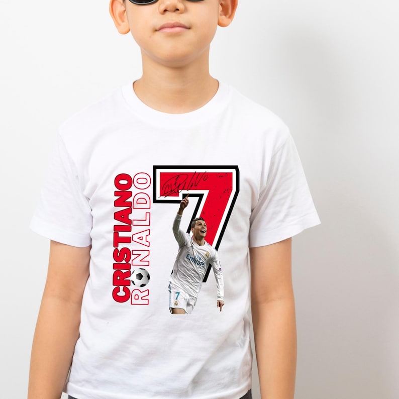 May include: White t-shirt featuring a graphic of soccer player Cristiano Ronaldo. The design includes the number 7 in red with a black outline, the player's name, and a soccer ball.