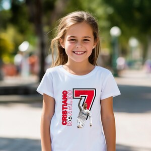 May include: A white t-shirt featuring a graphic of Cristiano Ronaldo in a soccer uniform, with the number 7 in red and black. The text "Cristiano Ronaldo" is printed vertically in red. A young girl is wearing the shirt.