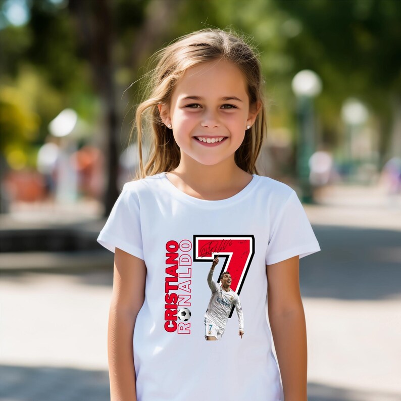 May include: A white t-shirt featuring a graphic of Cristiano Ronaldo in a soccer uniform, with the number 7 in red and black. The text "Cristiano Ronaldo" is printed vertically in red. A young girl is wearing the shirt.