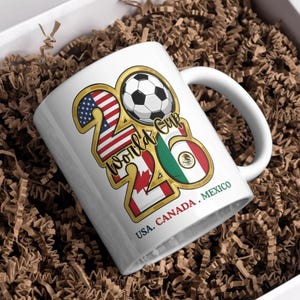 May include: A white ceramic mug with a black handle and the text "But First, Coffee" in a bold, black font. The mug is set against a blurred background, suggesting a cozy setting. The mug appears to be approximately 4 inches tall.