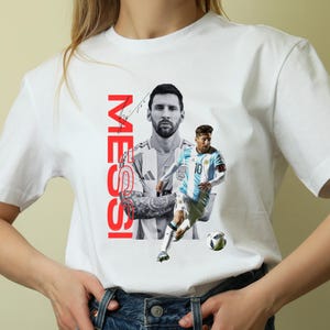 May include: White t-shirt featuring a graphic of a soccer player in a blue and white uniform, with the name "MESSI" in red vertical text. The design includes a black and white portrait of the player.