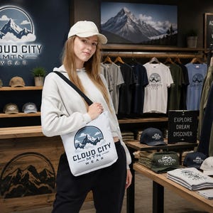 May include: A white tote bag featuring a circular logo with a mountain range and the text CLOUD CITY MTN WEAR. The bag has a black shoulder strap and is displayed in a retail store setting with various apparel and hats on shelves in the background.