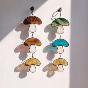 May include: Two vertical stained glass wall hangings, each featuring three mushroom shapes connected by metal rings. The left piece has brown and white glass, while the right piece displays green, blue, yellow, and white glass with dark metal solder lines.