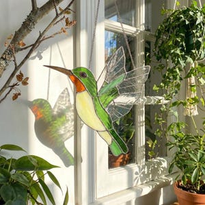 Op de afbeelding: Een gebrandschilderde kolibrie in groen, geel en helder hangt aan een ketting. De vogel heeft een oranje keel en een lange, puntige snavel. Zonlicht schijnt door het glas en werpt een kleurrijke schaduw op het raam.