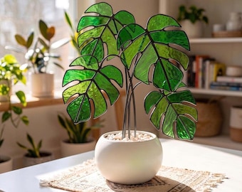 May include: A stained glass monstera plant sculpture featuring multiple vibrant green leaves with dark soldered seams. The glass leaves are supported by thin metal stems anchored in a smooth, round white ceramic pot filled with small white stones.