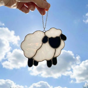 Könnte beinhalten: Ein Glasfenster-Sonnfänger in Form eines Schafs mit weißem Körper, schwarzem Gesicht und schwarzen Beinen. Das Schaf wird an einer silbernen Kette vor einem blauen Himmel mit weißen Wolken aufgehängt. Ein dekorativer Artikel.