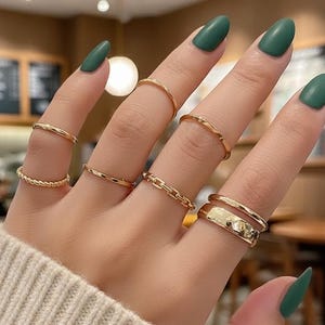 May include: A hand adorned with several gold rings in diverse styles, such as chain link, textured, and band designs. The hand features long, green-painted nails and is wearing a cream-coloured sweater.